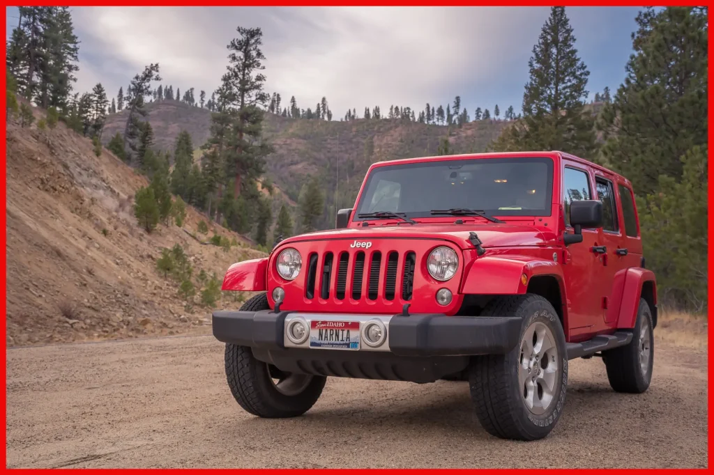 A red Jeep Wrangler parked on a dirt road with a forested mountainous backdrop under a cloudy sky.