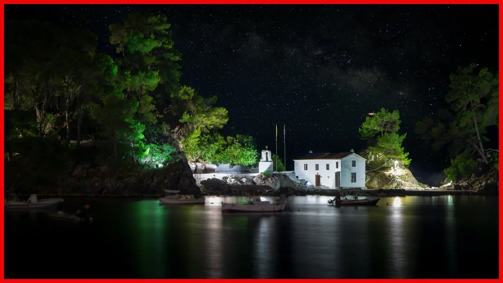 A night-time scene capturing a small white chapel nestled among lush evergreen trees on a secluded coast. The chapel and trees are illuminated with soft lighting against the backdrop of a magnificent starry sky. Small boats float gently on the calm waters, reflecting the twinkling stars above and the ambient lights from the shore.