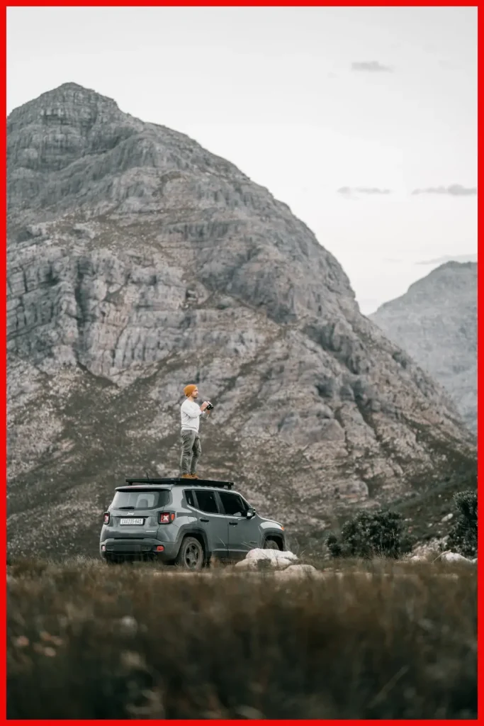 A person stands on a car roof with a camera in hand, capturing the vastness of the mountainous landscape that surrounds them.